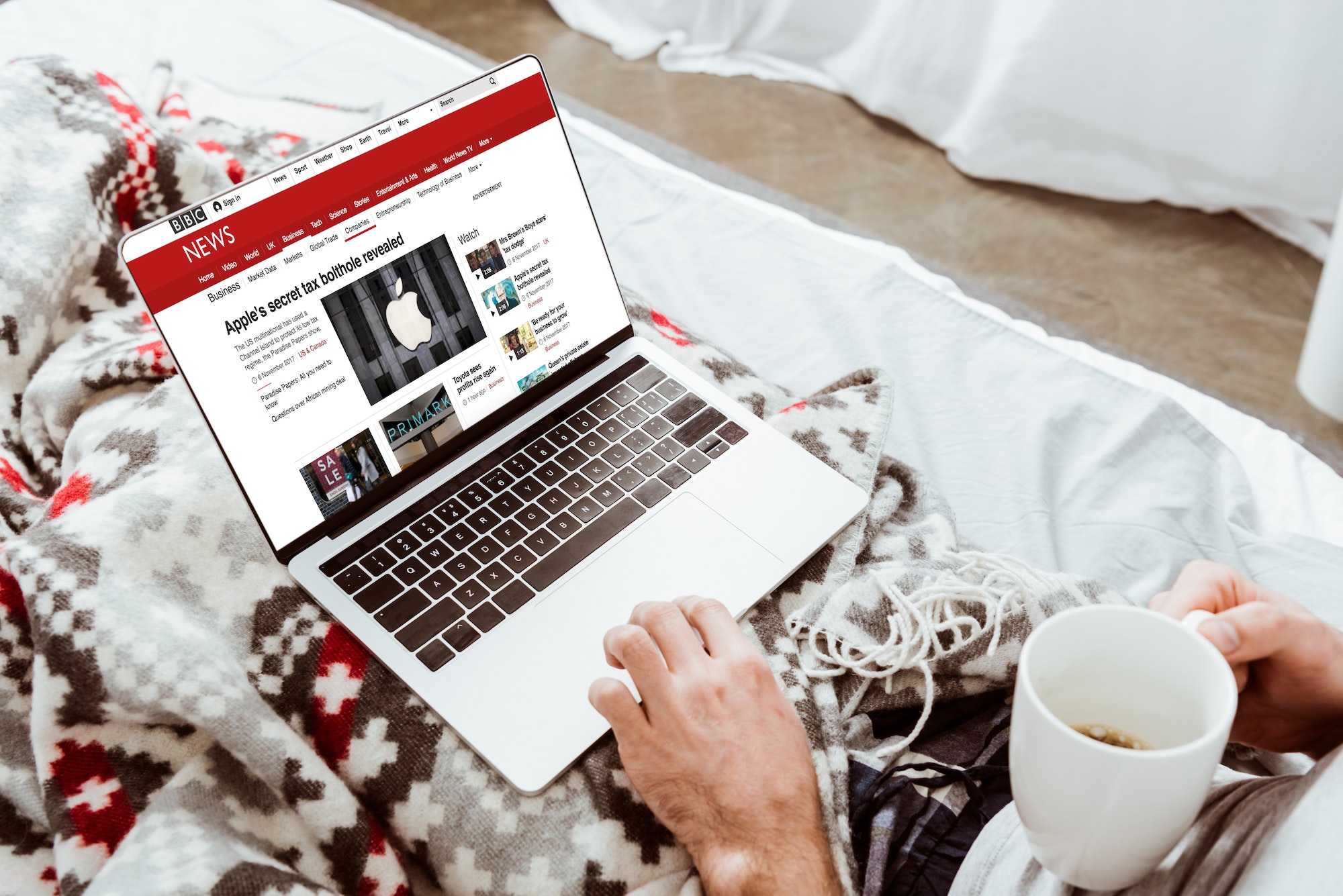 cropped image of man holding coffee cup and using laptop with bbc news on screen in bed at home